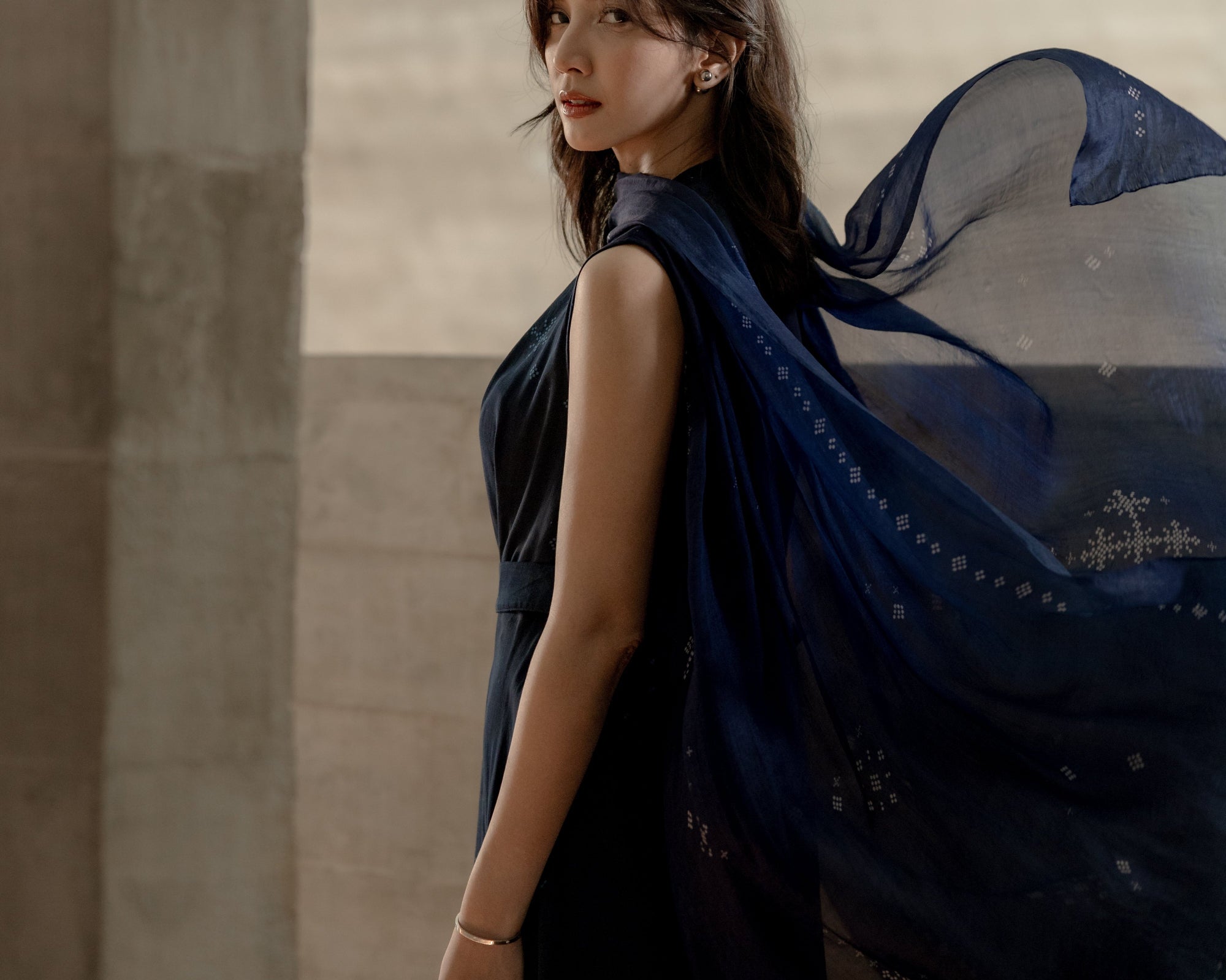 Sheila Dara wearing a dark ANGKASA Indigo dress with an exclusive scarf from SORE The Movie against a neutral wall.