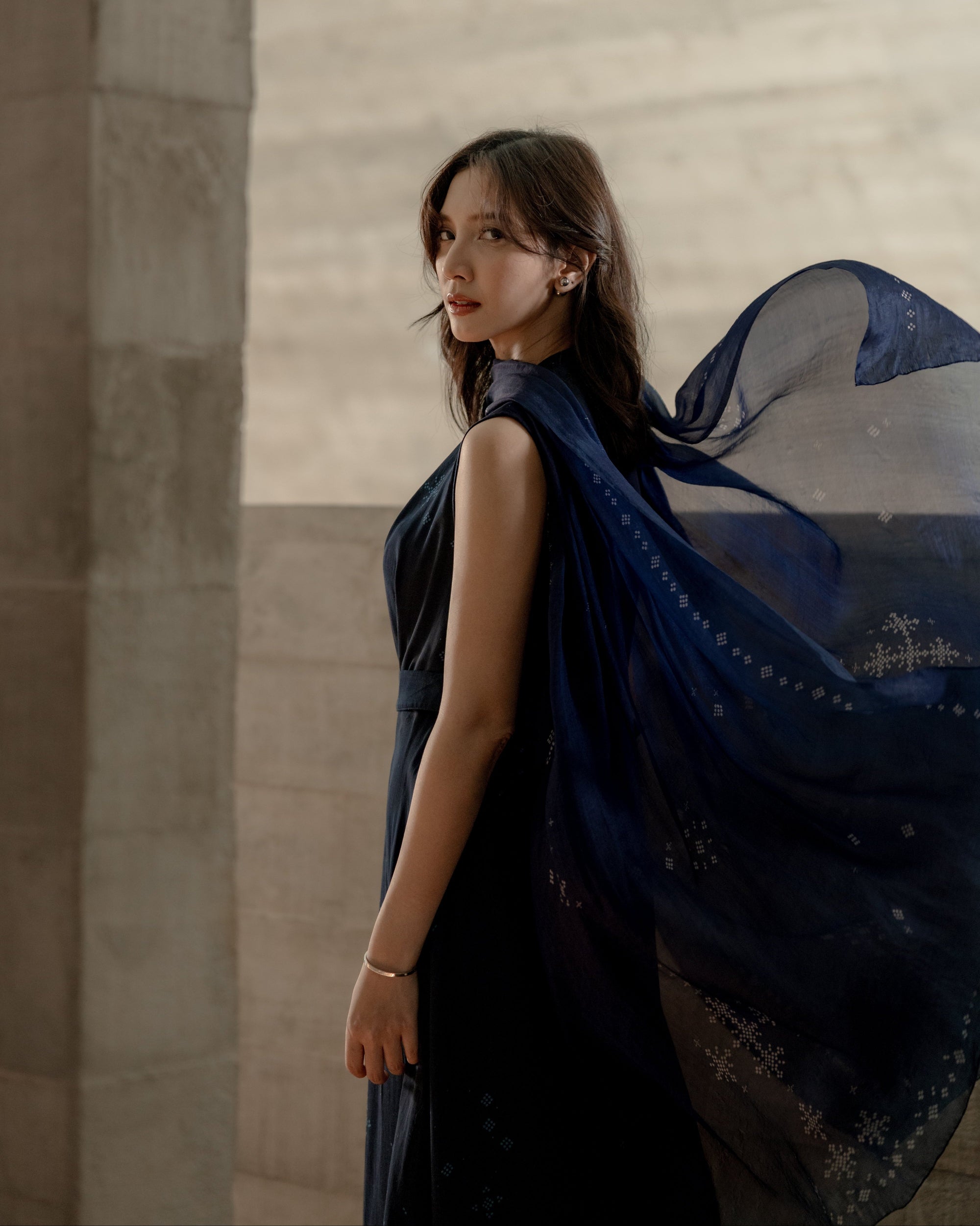 Sheila Dara wearing a dark ANGKASA Indigo dress with an exclusive scarf from SORE The Movie against a neutral wall.